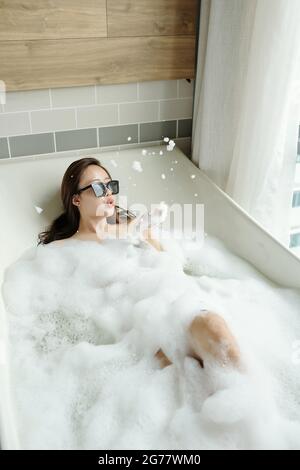 Yuong femme en lunettes de soleil couchée dans la salle de bains et jouant avec de la mousse Banque D'Images