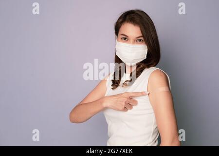 Femelle asiatique vaccinée montrant un bandage en plâtre sur le bras après avoir reçu l'injection du vaccin Covid-19 et sourire tout en étant assise portant un visage d'hygiène médicale M. Banque D'Images