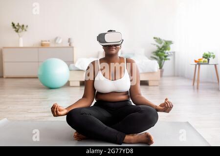 Femme noire de taille plus dans un casque VR, assise dans une posture lotus, méditant ou pratiquant le yoga à l'intérieur Banque D'Images