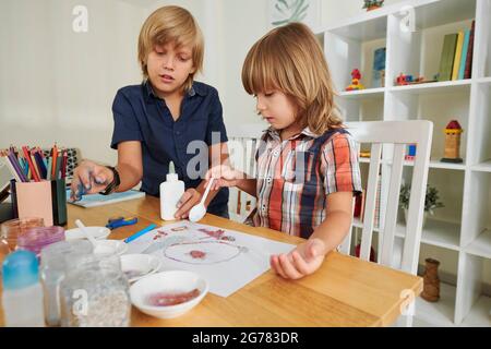 Garçon aidant frère cadet avec des paillettes encollantes sur feuille de papier quand ils apfinissant le temps ensemble à la maison Banque D'Images