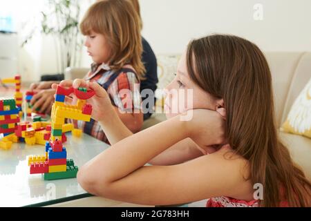 Une jeune fille pensive qui s'ennuie avec des blocs de jouets colorés et des tours de construction, ses frères jouant à proximité Banque D'Images