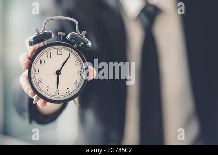 Main d'homme d'affaires en costume noir tenant et montrant l'horloge. La mise au point sélective laisse l'effet de flou en arrière-plan. Idée de valeur du temps dans le travail d'entreprise. Banque D'Images