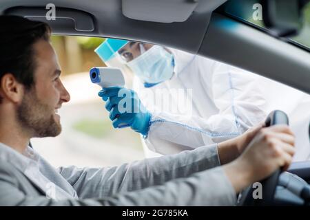 Le personnel médical en costume de noisette prend la température pour la fièvre à l'homme avant le test Covid-19 au poste de conduite à l'hôpital Banque D'Images