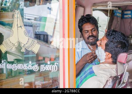 Indian barber brandising rasoir de gorge découpé donne à un client un rasage avec salutation Namaste sur porte en verre, tamil Nadu, Inde Banque D'Images