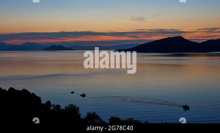 Iles Ioniennes, Ithaca, Molos Bay, Vathi, Dawn, Vue sur la baie de Molos, l'îlot de Skartsoubonisi et le continent et face à la baie, bateau de pêche crée des eaux dans la mer calme, ciel bleu à rouge, nuages gris Banque D'Images