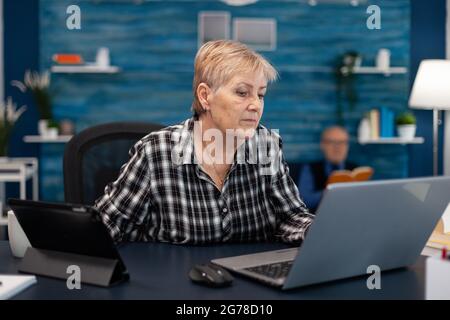 Propriétaire d'entreprise senior travaillant à domicile à l'aide d'un ordinateur portable. Femme âgée dans le salon à la maison utilisant une tablette pc portable de moder technoloy pour la communication assis au bureau à l'intérieur. Banque D'Images