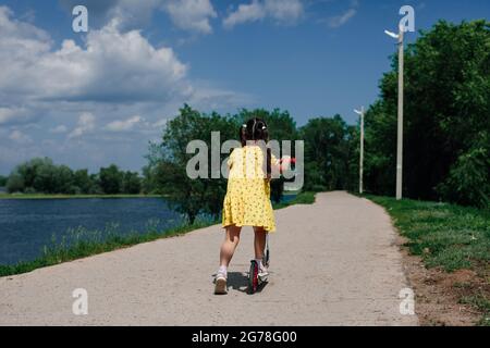 vue arrière d'une fille dans une robe jaune, une fille apprend à monter un scooter dans un parc naturel près du lac Banque D'Images