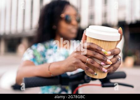 Une femme africaine non reconnue avec ses ongles a fait tenir une tasse de café tout en se tenant dans son scooter électrique. Banque D'Images