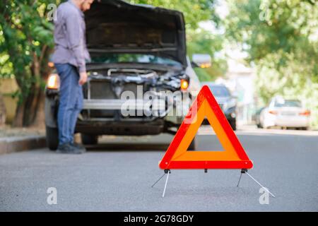 Triangle d'avertissement rouge d'accident de la route. Triangle devant la voiture de naufrage et le conducteur. La personne blessée en accident regarde sous le Banque D'Images