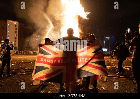 Belfast, Royaume-Uni. 11 juillet 2021. Un homme drape l'Union Jack autour de ses épaules devant un pyre flamboyant à la construction de feu de camp de Sandy Row dans le sud de Belfast.le 11 juillet ou la « onzième nuit » voit une nuit de feux de camp qui sont illuminés à minuit autour du nord de l'Irlande pour célébrer le tournant du 12 juillet. Cette année marque le centenaire de la formation de l'Irlande du Nord et est aussi la première marche Orange autorisée depuis le début de la pandémie. Crédit : SOPA Images Limited/Alamy Live News Banque D'Images