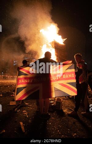 Belfast, Royaume-Uni. 11 juillet 2021. Un homme drape l'Union Jack autour de ses épaules devant un pyre flamboyant à la construction de feu de camp de Sandy Row dans le sud de Belfast.le 11 juillet ou la « onzième nuit » voit une nuit de feux de camp qui sont illuminés à minuit autour du nord de l'Irlande pour célébrer le tournant du 12 juillet. Cette année marque le centenaire de la formation de l'Irlande du Nord et est aussi la première marche Orange autorisée depuis le début de la pandémie. Crédit : SOPA Images Limited/Alamy Live News Banque D'Images