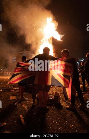 Belfast, Royaume-Uni. 11 juillet 2021. Un homme drape l'Union Jack autour de ses épaules devant un pyre flamboyant à la construction de feu de camp de Sandy Row dans le sud de Belfast.le 11 juillet ou la « onzième nuit » voit une nuit de feux de camp qui sont illuminés à minuit autour du nord de l'Irlande pour célébrer le tournant du 12 juillet. Cette année marque le centenaire de la formation de l'Irlande du Nord et est aussi la première marche Orange autorisée depuis le début de la pandémie. Crédit : SOPA Images Limited/Alamy Live News Banque D'Images