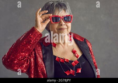 Portrait d'une belle femme âgée en lunettes de soleil rouges et veste à paillettes posé en studio Banque D'Images