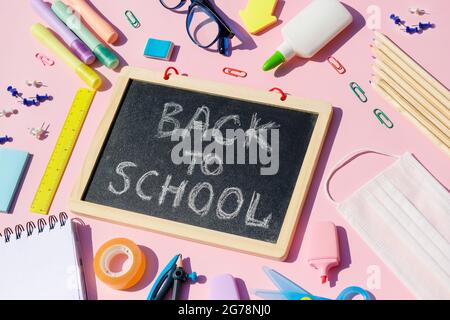 Retour à l'école avec fournitures scolaires, articles de papeterie, masque médical et carnet avec inscription de retour à l'école pour les enfants sur fond de bureau rose, dessus de vue plat. Banque D'Images