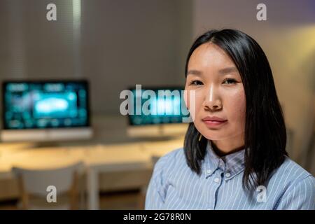 Portrait du contenu une jeune femme d'affaires asiatique confiante debout contre des ordinateurs dans un bureau sombre Banque D'Images