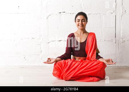 Jeune femme indienne adulte à sari méditant yoga lotus poser zen comme avec le signe ok mudra geste à la maison à l'intérieur. Banque D'Images