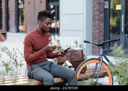 Travailleur africain sérieux de collier blanc en train de manger un sandwich et d'utiliser un smartphone Banque D'Images