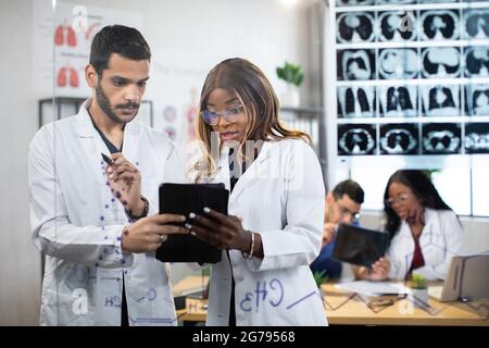 Deux médecins multiethniques, hommes et femmes, debout derrière un mur de verre avec IRM et travaillant sur un Tablet pc. Leurs collègues se parlent à la table en arrière-plan Banque D'Images