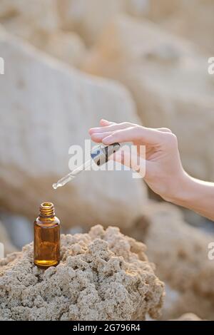 Pot cosmétique avec crème, sérum ou huile essentielle bio pour le visage et le corps, sur fond de pierres naturelles à la lumière du coucher du soleil. Salon de beauté et cosmétiques naturels. Main tenir compte-gouttes. Banque D'Images