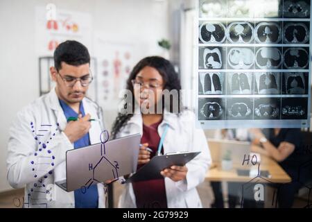 Vue rapprochée de deux scientifiques spécialisés et multiethniques, se tenant près de la paroi en verre avec des formules chimiques et une tomodensitométrie, à l'aide d'un ordinateur portable. Concentrez-vous sur le mur de verre Banque D'Images