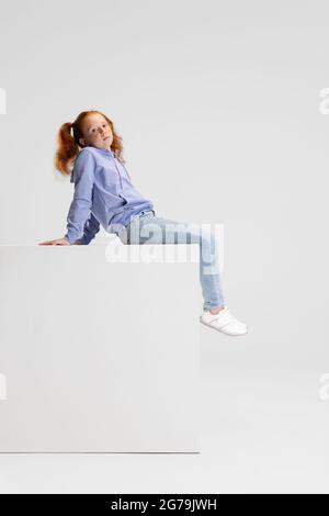 Une jolie fille à tête rouge dans des vêtements décontractés assis sur une grande boîte isolée sur fond blanc de studio. Concept d'enfance heureuse. Enfant ensoleillé. Semble heureux Banque D'Images