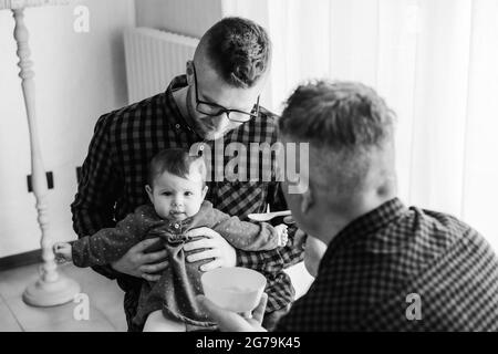 Homme gay couple avec adopté bébé fille à la maison - Deux beaux pères nourrissent la petite fille dans la cuisine - Baby-sitters masculins - famille LGBT à la maison - diversité Banque D'Images