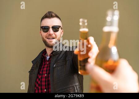 Un homme froid qui toaste de la bière avec le meilleur ami en plein air contre Un fond de mur vert - mains de l'homme branché portant veste en cuir brunissage avec be Banque D'Images
