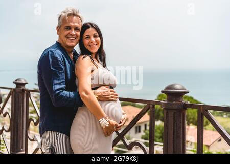Couple heureux avec femme enceinte debout contre un beau paysage de mer pendant les vacances d'été - jeune femme et mari riant ensemble expe Banque D'Images