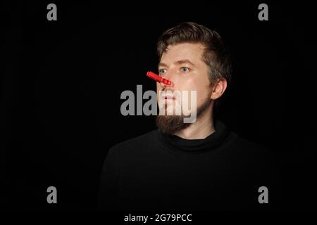 Homme barbu avec une épingle à linge sur le nez. Mauvaise piqûre, drôle de prankster, concept de plaisanterie. Rire de vous-même. Épinglette rouge - symbole de la moquerie et de la plaisanterie Banque D'Images