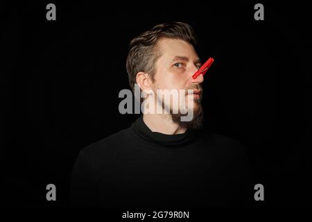 Homme barbu avec une épingle à linge sur le nez. Mauvaise piqûre, drôle de prankster, concept de plaisanterie. Rire de vous-même. Épinglette rouge - symbole de la moquerie et de la plaisanterie Banque D'Images