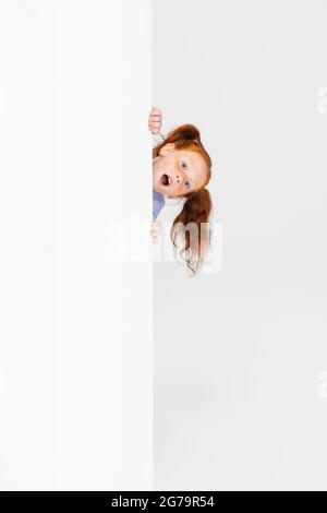 Portrait d'une adorable fille à tête rouge à revers dans un ensemble décontracté qui se pose sur le mur isolé sur fond blanc de studio. Concept d'enfance heureuse. Ensoleillé Banque D'Images