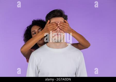 Une femme afro-américaine ferme les yeux de son ami bien-aimé avant du surprendre. Couple interracial sur fond de studio violet. Amour, vacances Banque D'Images