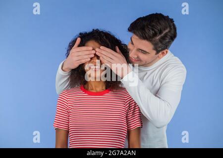 White man ferme les yeux de sa chère amie afro-américaine avant de la surprendre. Couple interracial sur fond de studio bleu. Amour, vacances Banque D'Images