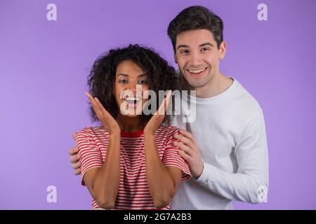 Homme blanc avec sa chère amie afro-américaine après la surprise. Couple interracial sur fond de studio violet. Amour, vacances, bonheur Banque D'Images