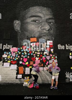 Ruby Swave, 7 ans (à gauche) et sa sœur Reeva Swave, 4 ans, placent des messages de soutien au-dessus des sacs poubelle qui ont été placés sur des mots offensifs sur la fresque de l'attaquant de Manchester United et du joueur d'Angleterre Marcus Rashford sur le mur du café Coffee House sur Copson Street, Withington, Qui semblait vandalisé le matin après que l'équipe de football d'Angleterre a perdu la finale de l'UEFA Euro 2021. Date de la photo: Lundi 12 juillet 2021. Banque D'Images