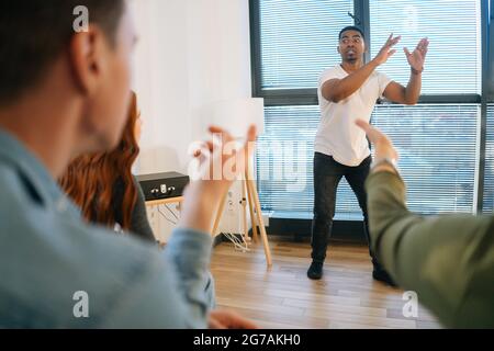 Portrait d'un homme afro-américain joyeux jouant des charades avec des amis montrant du pantomime par fenêtre dans une salle de bureau légère. Banque D'Images