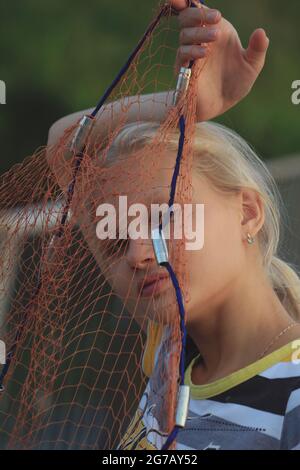 Portrait d'une jeune fille blonde à travers un filet de pêche. Blonde fille tenant des articles de pêche à l'extérieur devant son visage. Banque D'Images