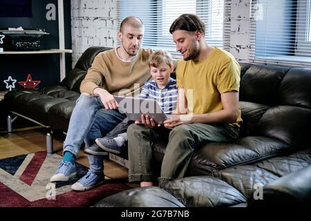 Deux jeunes hommes et un petit garçon assis sur un canapé à la maison et regardant des dessins animés ou des vidéos éducatives sur un ordinateur portable Banque D'Images