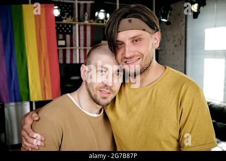 Portrait de deux hommes gais heureux qui s'embrasent en se tenant contre le drapeau arc-en-ciel et en regardant l'appareil photo Banque D'Images