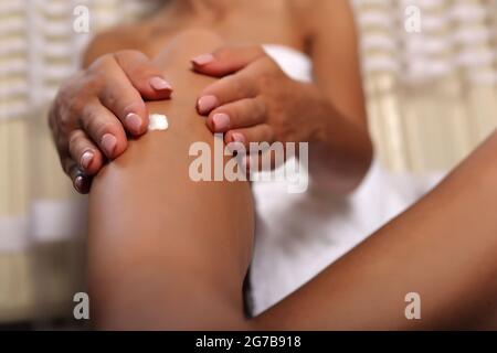 Femme mains appliquant de la crème pour le corps sur sa jambe à la maison. La fille porte une serviette blanche. Soin de la peau et concept de beauté Banque D'Images