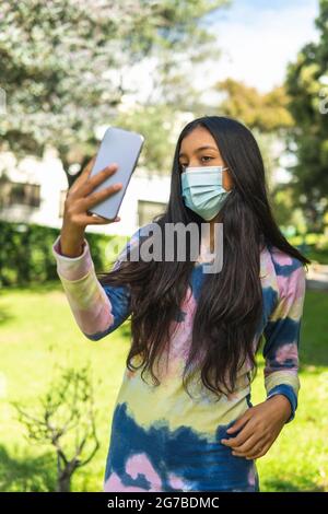 Long cheveux droits brunette Latina jeune fille avec masque prendre une photo de selfie avec son téléphone cellulaire à l'extérieur Banque D'Images