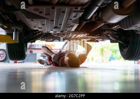 Mécanicien en uniforme couché et travaillant sous la voiture au garage d'entretien automobile, mécanicien de voiture réglant la tension dans l'élément de suspension du véhicule à auto repa Banque D'Images