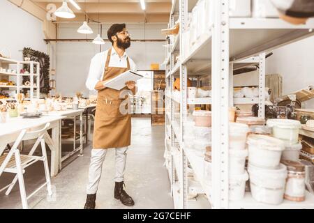 Jeune homme d'affaires séduisant un potter avec une barbe et une moustache travaille dans son atelier. Conserve les enregistrements et transcrites dans un ordinateur portable en inspectant Banque D'Images