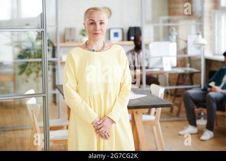 Portrait d'une femme d'affaires mûre avec des cheveux courts et dans une élégante robe jaune regardant l'appareil photo tout en se tenant au bureau Banque D'Images