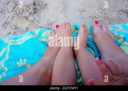 Gros plan des pieds de bébé assis entre le pied de la mère sur une serviette bleue sur le sable Banque D'Images