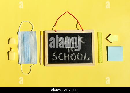 Fournitures de papeterie pour l'école, masque médical sur fond jaune, distanciation sociale, réouverture de l'école après une pandémie de covid-19. Nouveau concept normal.espace de copie de la vue de dessus,tableau noir. Banque D'Images