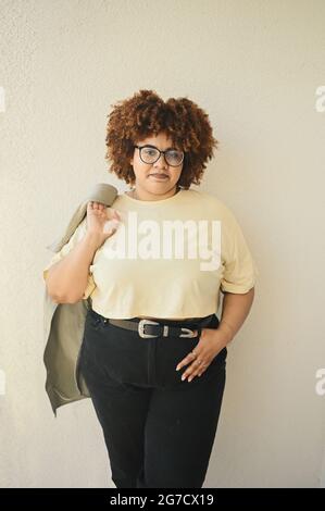 Beau sourire heureux curvy plus taille africaine noire femme cheveux afro posant dans le t-shirt beige, jeans, lunettes stylées sur fond beige. Corps imperfection, acceptation corps positif concept de diversité Banque D'Images