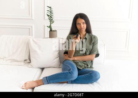 Jeune femme indienne avec télécommande regardant une série de télé ennuyeuse, en train de regarder malheureuse et fatiguée, femme de race mixte regardant la télévision dans le salon à la maison Banque D'Images