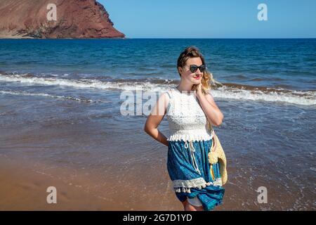 Jeune femme millénaire portant une robe à motif cachemire bleu et blanc, un sac de plage et des lunettes de soleil, souriant et appréciant la plage de la Tejita près de Montana Roja Banque D'Images