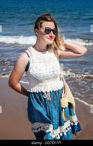 Jeune femme millénaire portant une robe à motif cachemire bleu et blanc, un sac de plage et des lunettes de soleil tout en profitant de vacances d'été à la plage et de vacances Banque D'Images